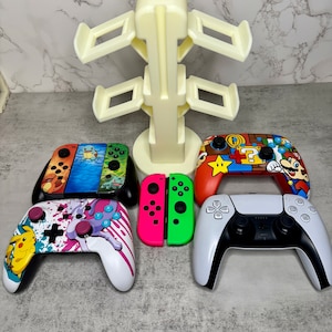 May include: A white plastic stand with four slots for holding video game controllers. Five controllers are shown: a white and pink controller with a Pikachu design, a red and green controller, a white controller with a Mario design, a blue and orange controller with a Pokemon design, and a white and black PlayStation 5 controller.