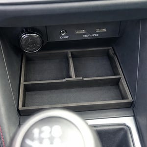 May include: A dark gray wooden car console organizer with multiple compartments. The organizer fits into the center console area of a vehicle. The car's interior features black leather with red stitching and a silver gear shift.