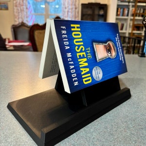 Puede incluir: Un soporte de libro negro con un libro azul titulado "The Housemaid" de Freida McFadden. La portada del libro presenta una cerradura con un ojo mirando a través de ella. El libro es un best-seller del New York Times.