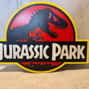 May include: A circular wall sign featuring the Jurassic Park logo with a black dinosaur silhouette against a red background, enclosed by a yellow border. The text JURASSIC PARK is displayed in white capital letters on a black rectangular base.