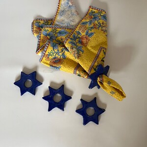 May include: A yellow floral patterned napkin with a blue napkin ring. Three additional blue star-shaped napkin rings are also visible. The napkin has a floral design with blue and orange accents.
