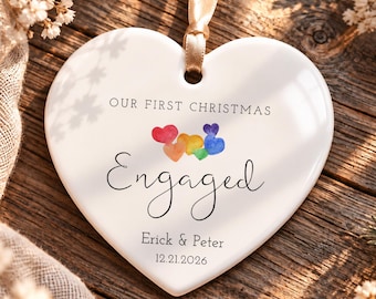 May include: A white heart-shaped ceramic ornament with a gold ribbon, featuring the text "OUR FIRST CHRISTMAS", "Engaged", "Erick & Peter", and "12.21.2026" printed in black, along with a small cluster of colorful watercolor hearts.