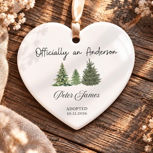 May include: A white heart-shaped ceramic ornament featuring a gold ribbon hanger. The text reads Officially an Anderson, Peter James, ADOPTED, and 10.11.2026. Three watercolor-style green pine trees are printed in the center of the ornament.
