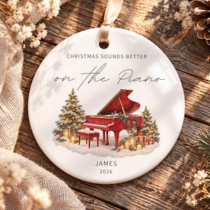 May include: A round white ceramic ornament with a gold ribbon hanger. The design features a red grand piano, snow-covered pine trees, and lanterns. Text reads: CHRISTMAS SOUNDS BETTER on the Piano, JAMES, 2026. The illustration includes festive greenery.