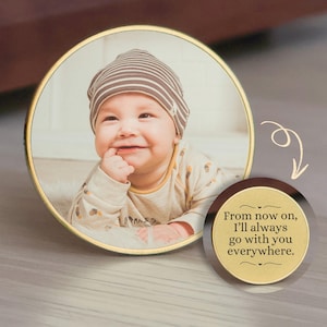 May include: A round photo frame with a gold rim displays a smiling baby wearing a striped hat and a light-colored onesie. A smaller, circular accent piece with text reads, "From now on, I'll always go with you everywhere."