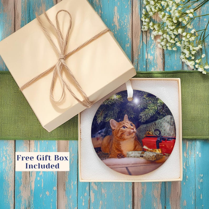May include: A round ceramic ornament featuring a ginger cat sitting under a Christmas tree with wrapped gifts. The ornament is presented in a square beige gift box with a tied twine bow. The text Free Gift Box Included is visible.