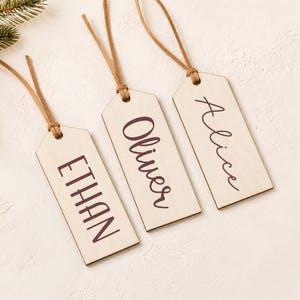 May include: Three rectangular light-colored wooden tags with rounded top corners, each featuring a brown leather cord loop. The tags are personalized with the names ETHAN, Oliver, and Alice printed in a dark brown font on the front surface.