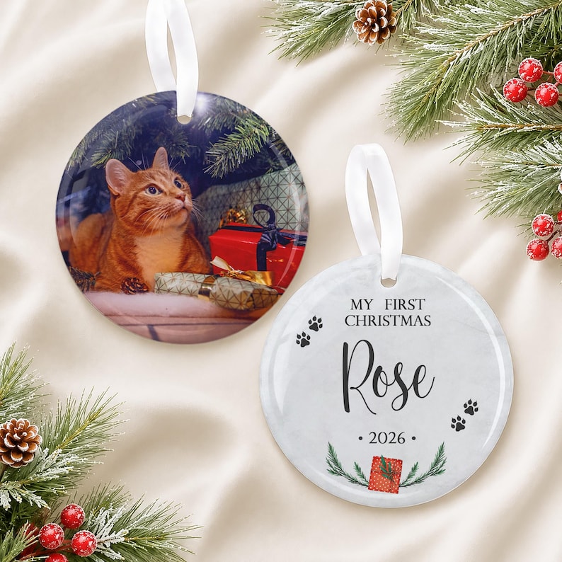 May include: Two round ceramic ornaments with white ribbon hangers. One ornament features a photograph of an orange tabby cat sitting near red and gold wrapped gifts. The other white ornament displays the text My First Christmas Rose 2026.
