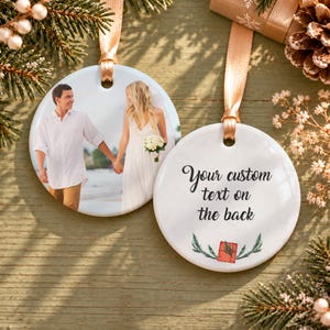 May include: Two round white ceramic ornaments with gold ribbon hangers. One ornament features a color photograph of a couple holding hands on a beach. The other ornament displays the black text Your custom text on the back above a small red gift icon.