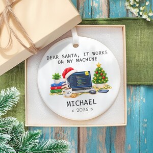 May include: A round white ceramic ornament featuring the text "DEAR SANTA, IT WORKS ON MY MACHINE" above a graphic of a laptop with a Santa hat, a small Christmas tree, books, and a mug. The name "MICHAEL" and "< 2026 >" are printed below.