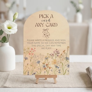 May include: Wooden sign with the text "PICK A card ANY CARD" and a floral design. The sign includes the text "PLEASE WRITE A MESSAGE AND SIGN YOUR NAME, SO WE CAN REMEMBER THIS SPECIAL DAY ANY TIME WE PLAY!" It is displayed on a small wooden easel.
