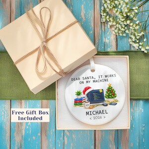 May include: A round white ceramic ornament featuring a graphic of a laptop with code, a Santa hat, a small Christmas tree, and books. The text reads: DEAR SANTA, IT WORKS ON MY MACHINE, MICHAEL, and 2026. The ornament is inside a square gift box.