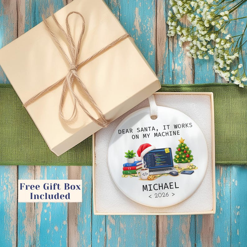 May include: A round white ceramic ornament featuring a graphic of a laptop with code, a Santa hat, a small Christmas tree, and books. The text reads: DEAR SANTA, IT WORKS ON MY MACHINE, MICHAEL, and 2026. The ornament is inside a square gift box.
