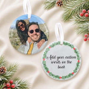 Puede incluir: Dos adornos redondos de cer&aacute;mica con cintas blancas. Uno muestra una fotograf&iacute;a en color de dos personas con gafas de sol al aire libre. El otro presenta un borde de corona de hojas verdes y el texto Add your custom words on the back.