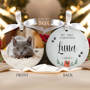 May include: Two round ceramic ornaments with white satin ribbons. The front features a gray kitten on a textured surface. The back displays the text MY FIRST CHRISTMAS, the name Luna, the year 2025, paw prints, and a small red gift box illustration.