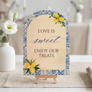 May include: Wooden sign with an arched top, featuring the text "LOVE IS sweet ENJOY OUR TREATS". The sign is decorated with a blue and white tile border and lemon illustrations. It is displayed on a small wooden easel.