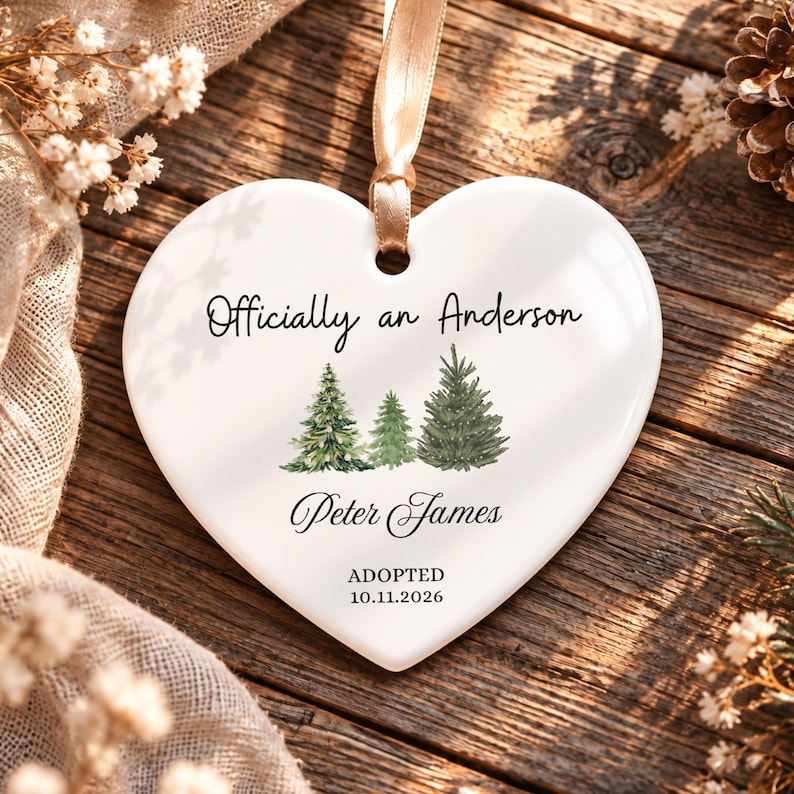 May include: A white heart-shaped ceramic ornament featuring a gold ribbon hanger. The text reads Officially an Anderson, Peter James, ADOPTED, and 10.11.2026. Three watercolor-style green pine trees are printed in the center of the ornament.