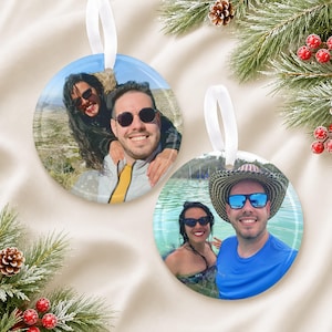 May include: Two round ceramic ornaments featuring custom photo prints. The first ornament displays a couple outdoors wearing sunglasses. The second ornament shows a couple in a body of water, with one wearing a wide-brimmed hat and sunglasses.