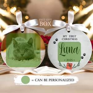 May include: Two round white ceramic ornaments with white satin ribbons. One features a green-tinted photo of a cat. The other displays the text MY FIRST CHRISTMAS, the name Luna, the year 2025, paw prints, and a small red gift box illustration.