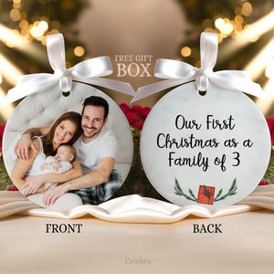 Puede incluir: Dos adornos redondos de cer&aacute;mica con cintas de sat&eacute;n blanco. El frente muestra una fotograf&iacute;a de una familia de tres. El reverso presenta el texto negro 'Our First Christmas as a Family of 3' sobre una peque&ntilde;a ilustraci&oacute;n de un regalo rojo.