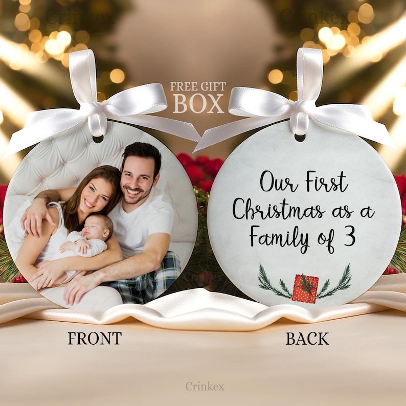 Puede incluir: Dos adornos redondos de cer&aacute;mica con cintas de sat&eacute;n blanco. El frente muestra una fotograf&iacute;a de una familia de tres. El reverso presenta el texto negro 'Our First Christmas as a Family of 3' sobre una peque&ntilde;a ilustraci&oacute;n de un regalo rojo.