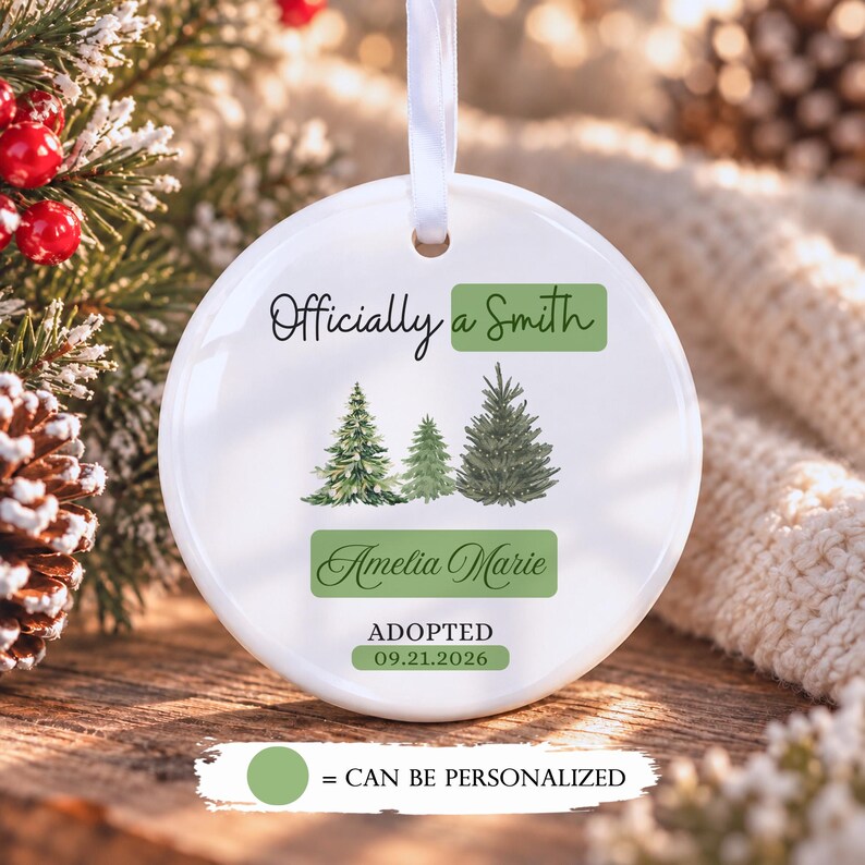 May include: A round white ceramic ornament with a white ribbon hanger. The text reads Officially a Smith, Amelia Marie, and ADOPTED 09.21.2026. It features three watercolor-style evergreen trees and green rectangular accents for personalization.