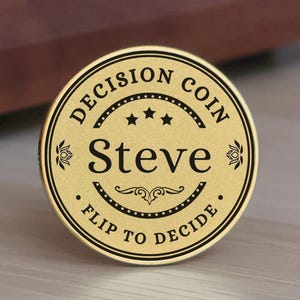 May include: A circular gold-toned metal decision coin featuring black engraved text that reads DECISION COIN at the top and FLIP TO DECIDE at the bottom, with the name Steve centered in large black lettering between three stars and a flourish.