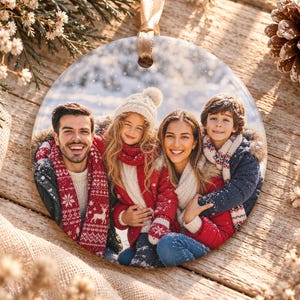 May include: A round ceramic ornament featuring a printed photograph of a family of four in a snowy outdoor setting. The family members are wearing winter clothing including red and white patterned scarves and sweaters with reindeer motifs.