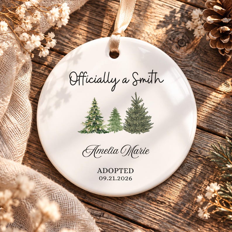May include: A round white ceramic ornament featuring a gold ribbon hanger. The text reads Officially a Smith, Amelia Marie, and ADOPTED 09.21.2026. Three watercolor-style green pine trees are printed in the center of the ornament.