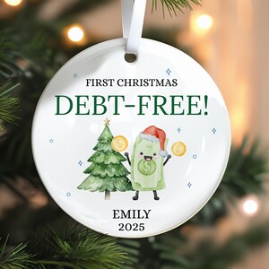 May include: White ceramic Christmas ornament with the text "FIRST CHRISTMAS DEBT-FREE!" and the name "EMILY 2025". The ornament features a cartoon dollar bill wearing a Santa hat, a Christmas tree, and gold coins.