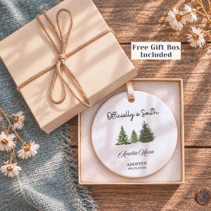 May include: A round white ceramic ornament featuring a watercolor illustration of three evergreen trees. The text reads Officially a Smith, Amelia Marie, and ADOPTED 09.21.2026. The ornament is presented in an open beige gift box with a twine bow.