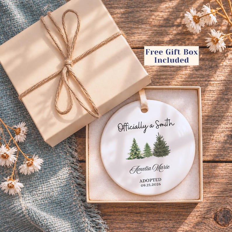 May include: A round white ceramic ornament featuring a watercolor illustration of three evergreen trees. The text reads Officially a Smith, Amelia Marie, and ADOPTED 09.21.2026. The ornament is presented in an open beige gift box with a twine bow.