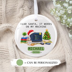 May include: A round white ceramic Christmas ornament featuring the text DEAR SANTA, IT WORKS ON MY MACHINE. The graphic shows a laptop with code, a Santa hat, a stack of books, a coffee mug, a Christmas tree, and the name MICHAEL with 2026.