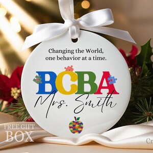 May include: White ceramic ornament with a white ribbon. The ornament has the text "Changing the World, one behavior at a time." and "BCBA Mrs. Smith" with a colorful puzzle piece apple.