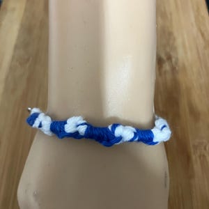 Peut inclure: Un bracelet noué bleu et blanc. Le bracelet est fait d'une matière tissée, avec des sections alternées de blanc et de bleu. Le bracelet est porté au poignet.
