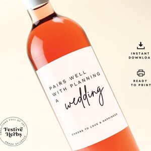 以下が含まれることがあります： 「Pairs well with planning a wedding」と「Cheers to love & happiness」の文字が書かれた白いワインボトルのラベル。ラベルはピンクのワインボトルに貼られています。