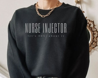 Könnte beinhalten: Ein schwarzer Pullover mit dem Aufdruck "NURSE INJECTOR let's talk about it" auf der Vorderseite. Das Model trägt den Pullover mit Jeans-Shorts.