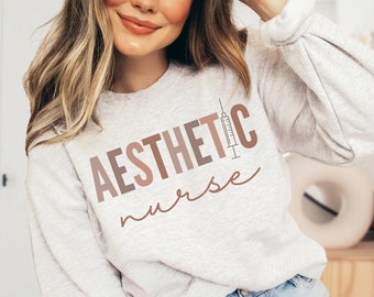 May include: A light gray sweatshirt with the text "AESTHETIC nurse" in brown and pink lettering. A stylized syringe is included in the text.