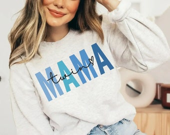 Puede incluir: Una sudadera gris con un diseño degradado azul y turquesa que dice "Mama twins" con un pequeño corazón.