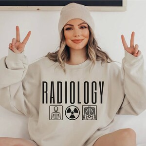 May include: A beige sweatshirt with the word "RADIOLOGY" printed in black. The sweatshirt has three black icons: a skeleton, a radiation symbol, and a skeleton with a spine.