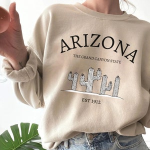 May include: A beige sweatshirt with a black graphic print of cacti and the text "Arizona, The Grand Canyon State, Est 1912."