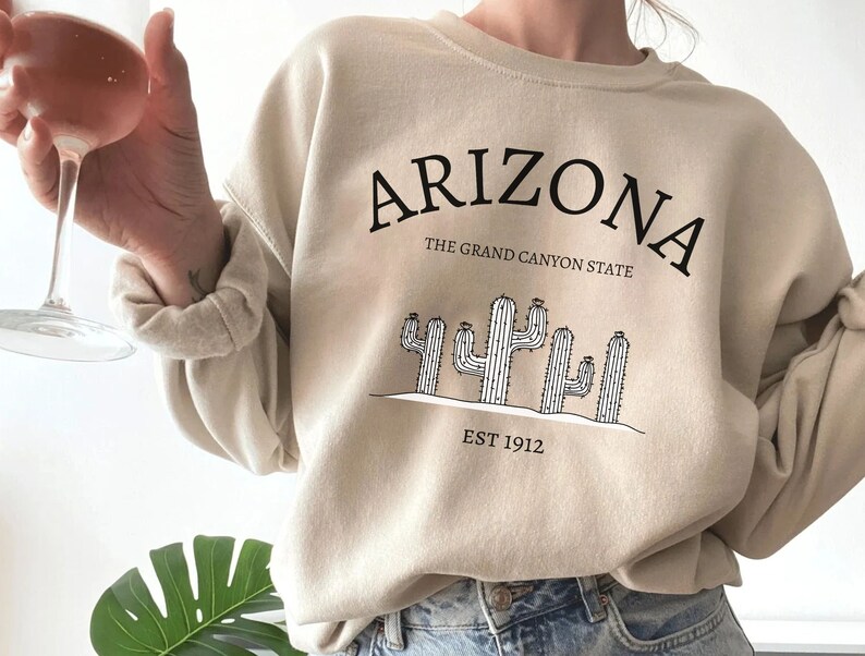 May include: A beige sweatshirt with a black graphic print of cacti and the text "Arizona, The Grand Canyon State, Est 1912."