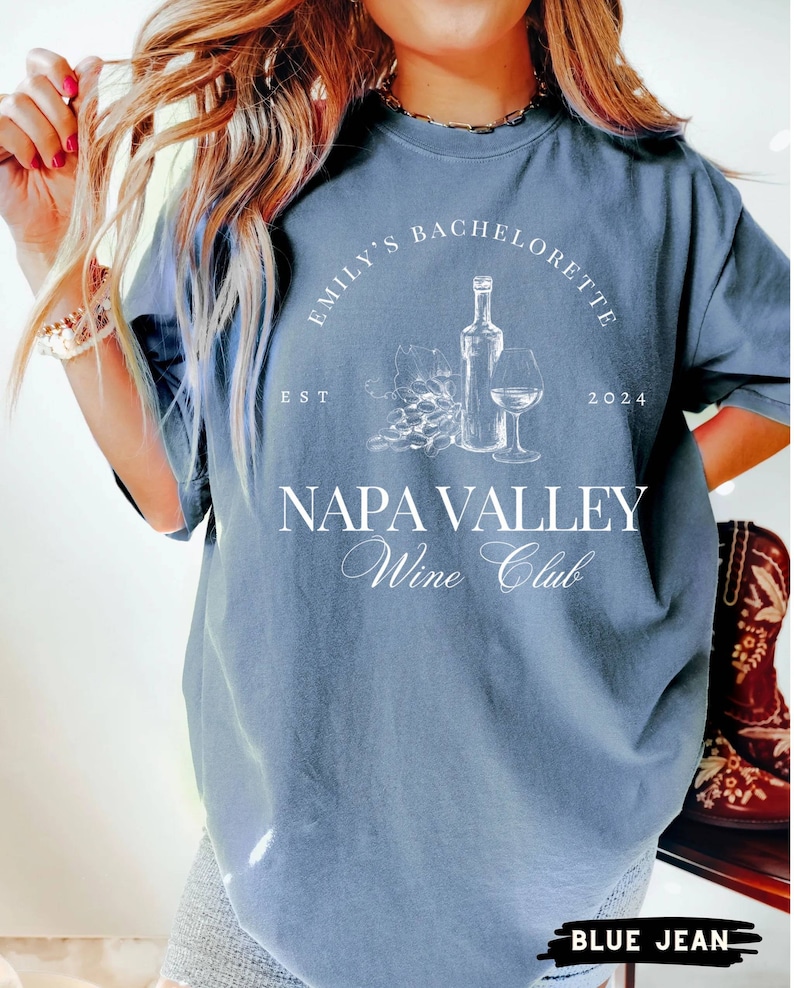 May include: A blue t-shirt with white text that reads "Emily's Bachelorette Est 2024 Napa Valley Wine Club". The shirt features a graphic of a wine bottle, grapes, and a wine glass.