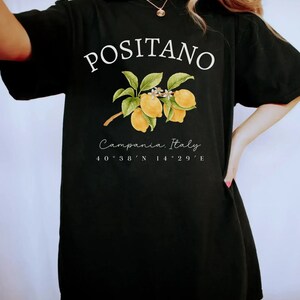 Może przedstawiać: Czarny t-shirt z białym napisem "POSITANO", z ilustracją gałązki cytryny. Poniżej znajduje się napis "Campania, Italy" oraz wsp&oacute;łrzędne 40&deg;38'N 14&deg;29'E.