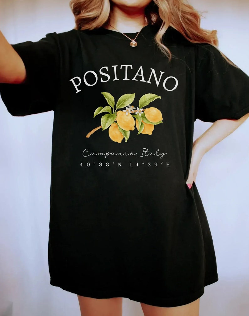 Może przedstawiać: Czarny t-shirt z białym napisem "POSITANO", z ilustracją gałązki cytryny. Poniżej znajduje się napis "Campania, Italy" oraz wsp&oacute;łrzędne 40&deg;38'N 14&deg;29'E.