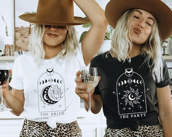 May include: Two women wearing white and black t-shirts with celestial designs and the phrases "THE BRIDE" and "THE PARTY". Both shirts feature a crescent moon, phases of the moon, and sun graphics. They are holding wine glasses and wearing brown hats.