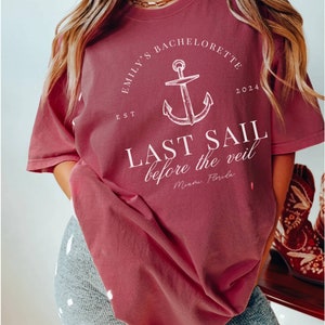 May include: A maroon t-shirt with a white anchor graphic and the text "Emily's Bachelorette Est 2024 Last Sail Before the Veil Miami Florida"