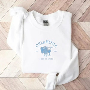 Puede incluir: Sudadera blanca con un estampado gr&aacute;fico azul de un b&uacute;falo y el texto "OKLAHOMA EST 1907 SOONER STATE".