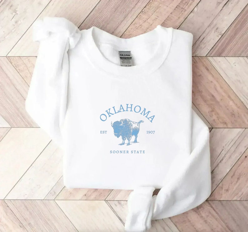 Puede incluir: Sudadera blanca con un estampado gr&aacute;fico azul de un b&uacute;falo y el texto "OKLAHOMA EST 1907 SOONER STATE".