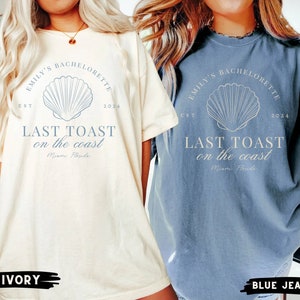 K&ouml;nnte beinhalten: Zwei &uuml;bergro&szlig;e T-Shirts mit einem Muschel-Motiv und dem Text "Emily's Bachelorette Est 2024 Last Toast on the Coast Miami Florida". Das Shirt links ist elfenbeinfarben und das Shirt rechts ist jeansblau.